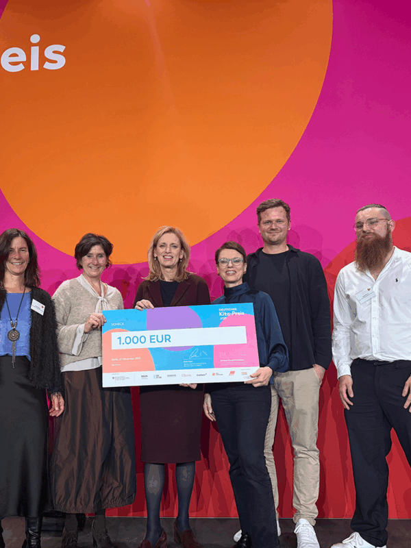 Das Team beim Kita-Preis Finale 2025 6 Personen stehen vor der bunten Kitapreis-Wand und halten einen großen Scheck in den Händen